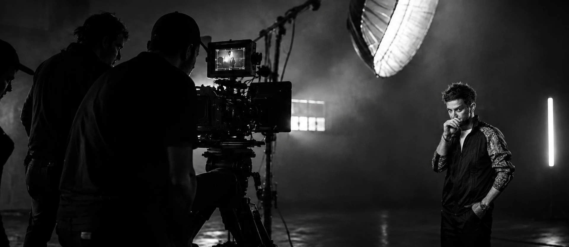 Black-and-white behind-the-scenes photo: a cinema camera and crew filming a subject on a smoke-filled set